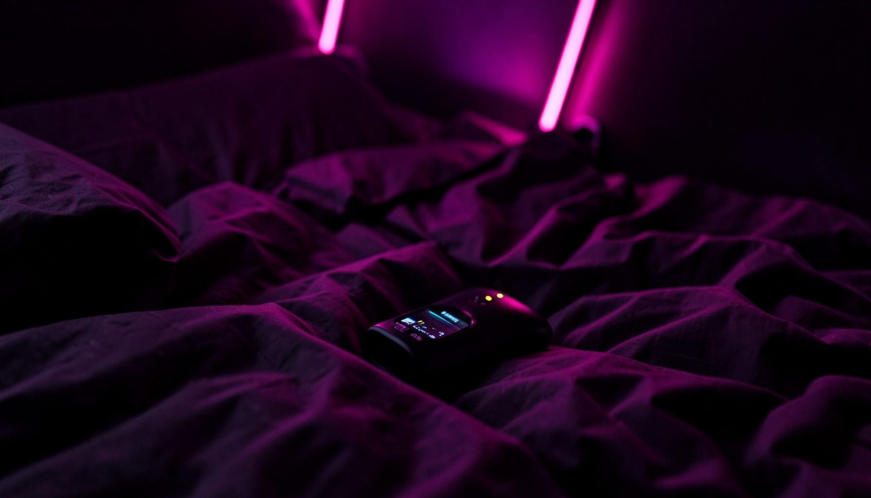 Dark bedroom with neon pink accents, a handheld device resting on rumpled sheets